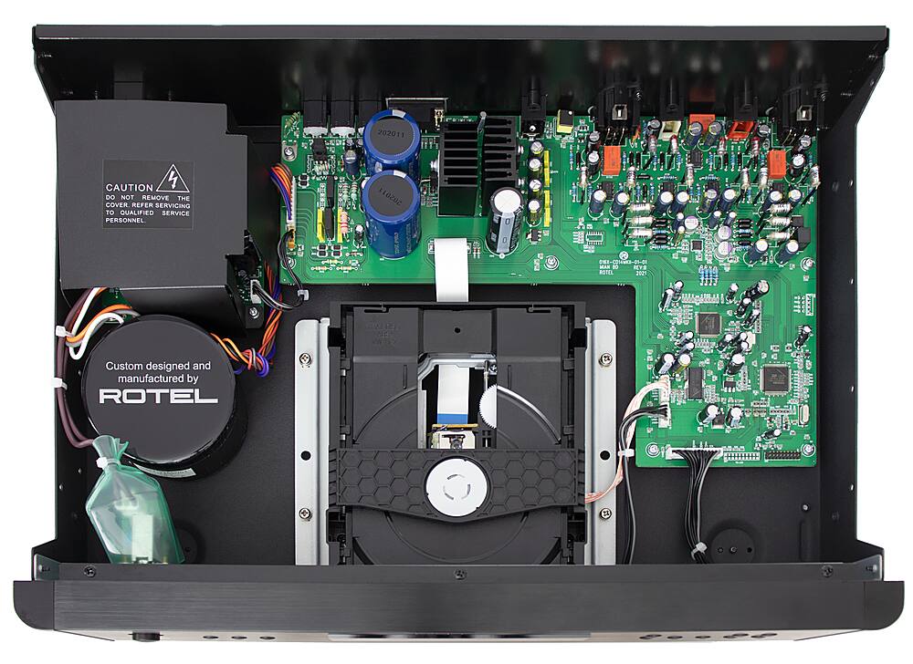 The image shows the inside of a device, likely a DVD player or a computer, with a green circuit board and various wires and components. The text on the image reads: "CAUTION DO NOT REMOVE THE COVER REFER SERVICING TO GUALIFIED SERVICE PERSONNEL 110V 0.1A 50/60Hz ROTEL 2021 1 E Custom designed and manufactured by ROTEL." This text indicates that the device should not be disassembled or tampered with by unqualified personnel, and that it is designed and manufactured by the ROTEL company.