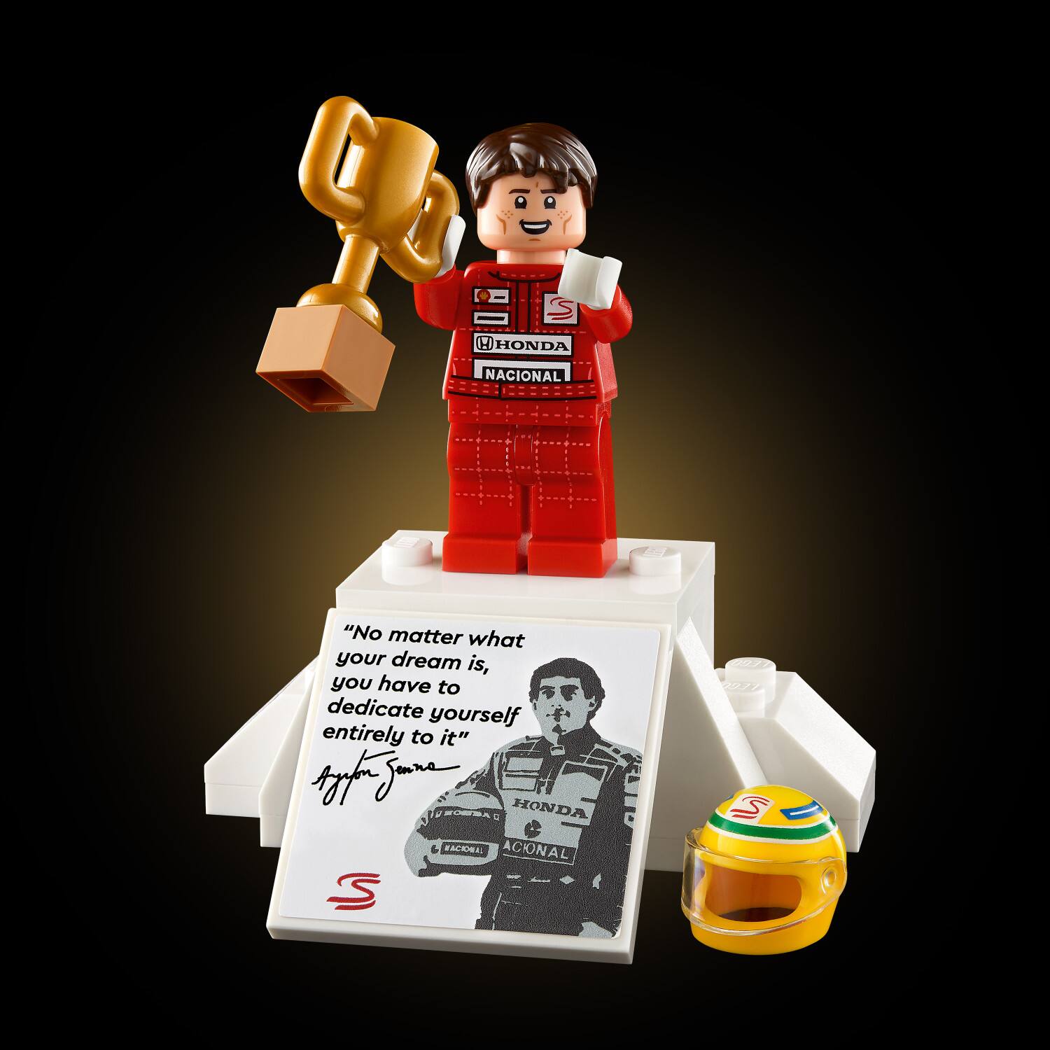 The image features a Lego figure holding a trophy and a cup, with a quote displayed nearby. The quote reads, "No matter what your dream is, you have to dedicate yourself entirely to it." The scene is set in a dark background, and there is a small sign with a picture of a man on it. Additionally, there is a small car and a book in the scene.