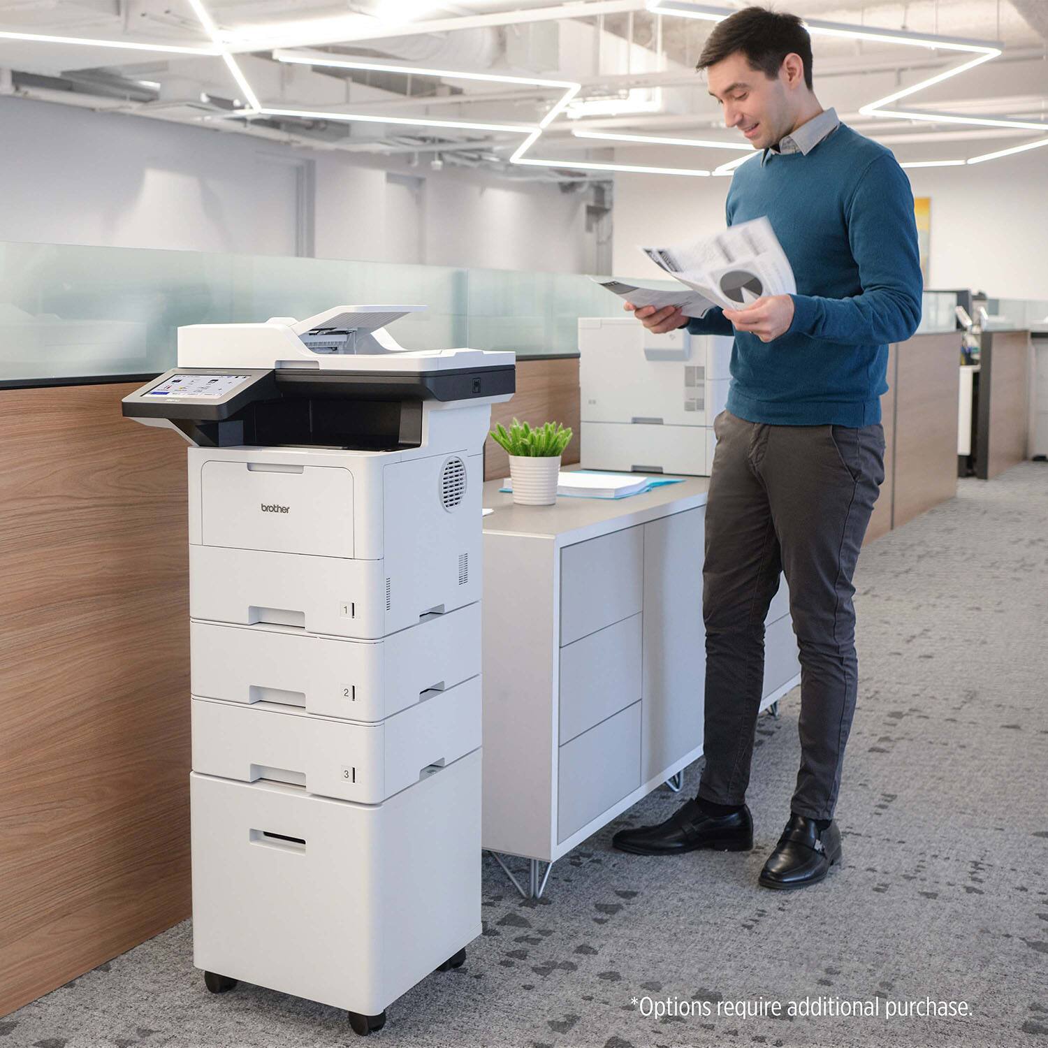 The image features a man standing in an office next to a Brother printer. The printer is white and has a cup placed on it. The man is holding a book, possibly reading or referencing it. The office appears to be well-equipped with multiple printers and a potted plant, creating a professional environment.