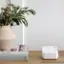The image features a white vase filled with green leaves and flowers, placed on a table. Next to the vase, there is a book and a remote control, which is likely used for a security system. The remote control has a cellular LTE backup feature, which requires the Arlo Safe & Secure Pro plan. The table is also accompanied by a small white device, possibly a sensor or a hub for the security system.