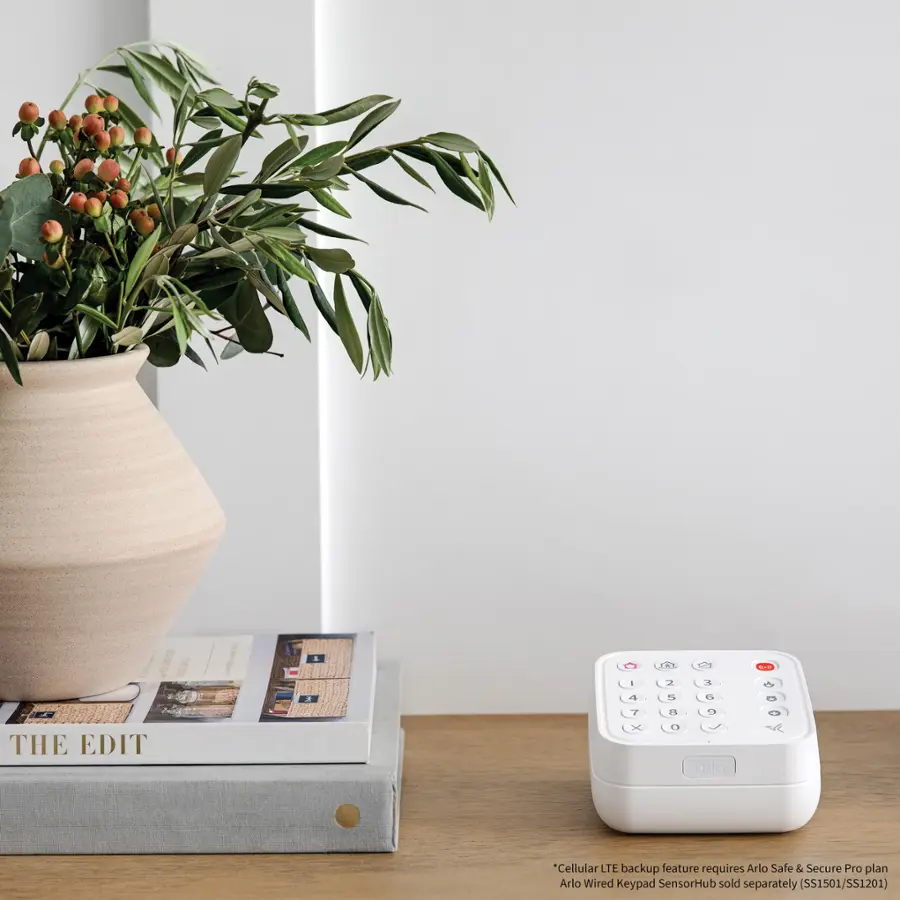 The image features a vase with a plant in it, placed on a table. The table also has a book and a small device, possibly a remote or a security system, next to the vase. The scene appears to be a cozy living space with a touch of greenery from the plant.