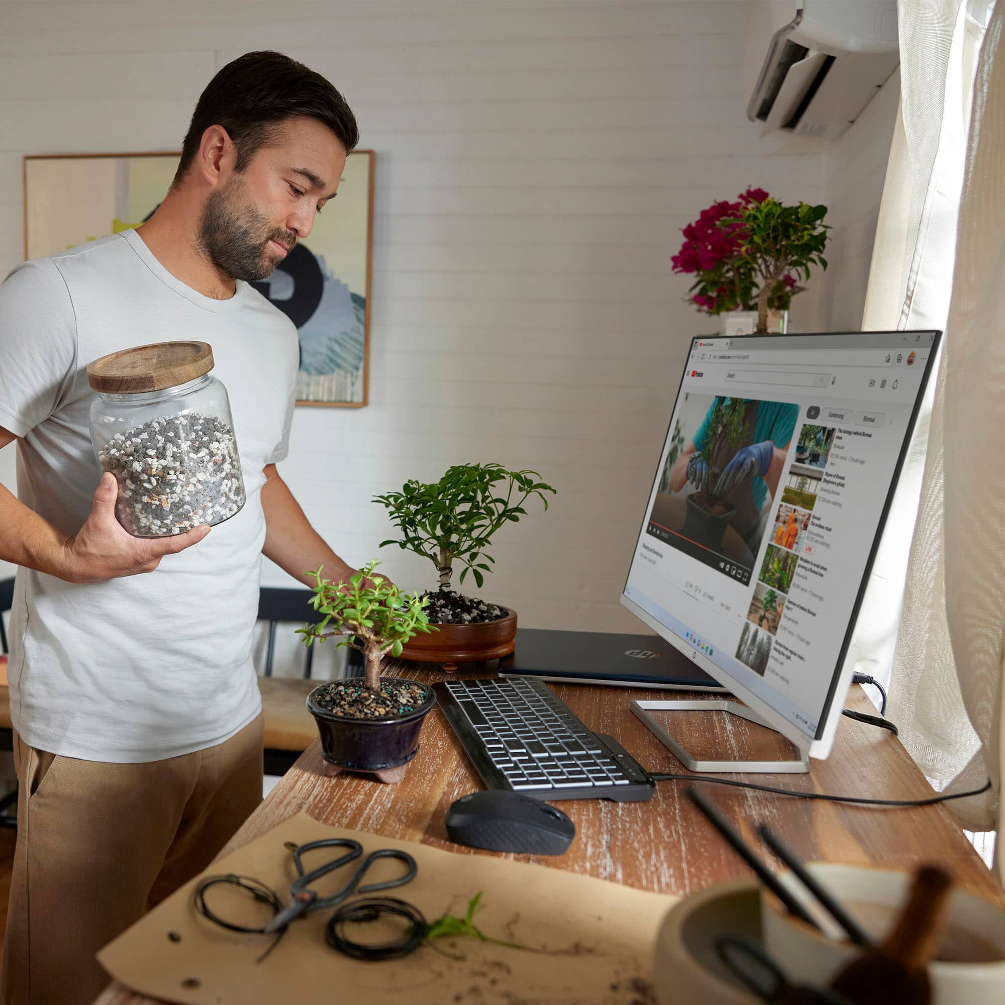 A man is standing in front of a computer with a potted plant and a jar of beads. He is holding the jar of beads in one hand and the potted plant in the other. There is a keyboard on the desk in front of the computer, and a laptop is also present. The man appears to be focused on the computer screen, possibly working or browsing the internet.