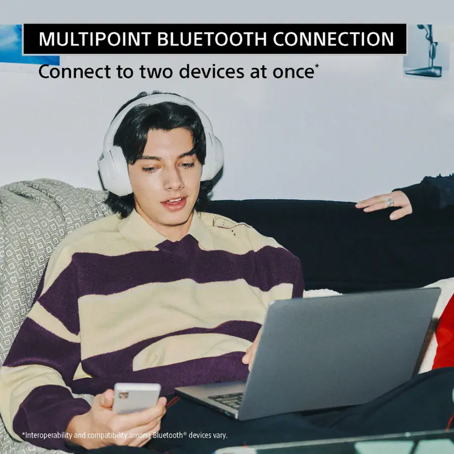 The image features a man sitting on a couch, wearing headphones and using a laptop. The man is also holding a cell phone in his hand. The scene suggests that the man is using a multipoint Bluetooth connection, which allows him to connect to two devices at once. However, the compatibility and interoperability of Bluetooth devices may vary.