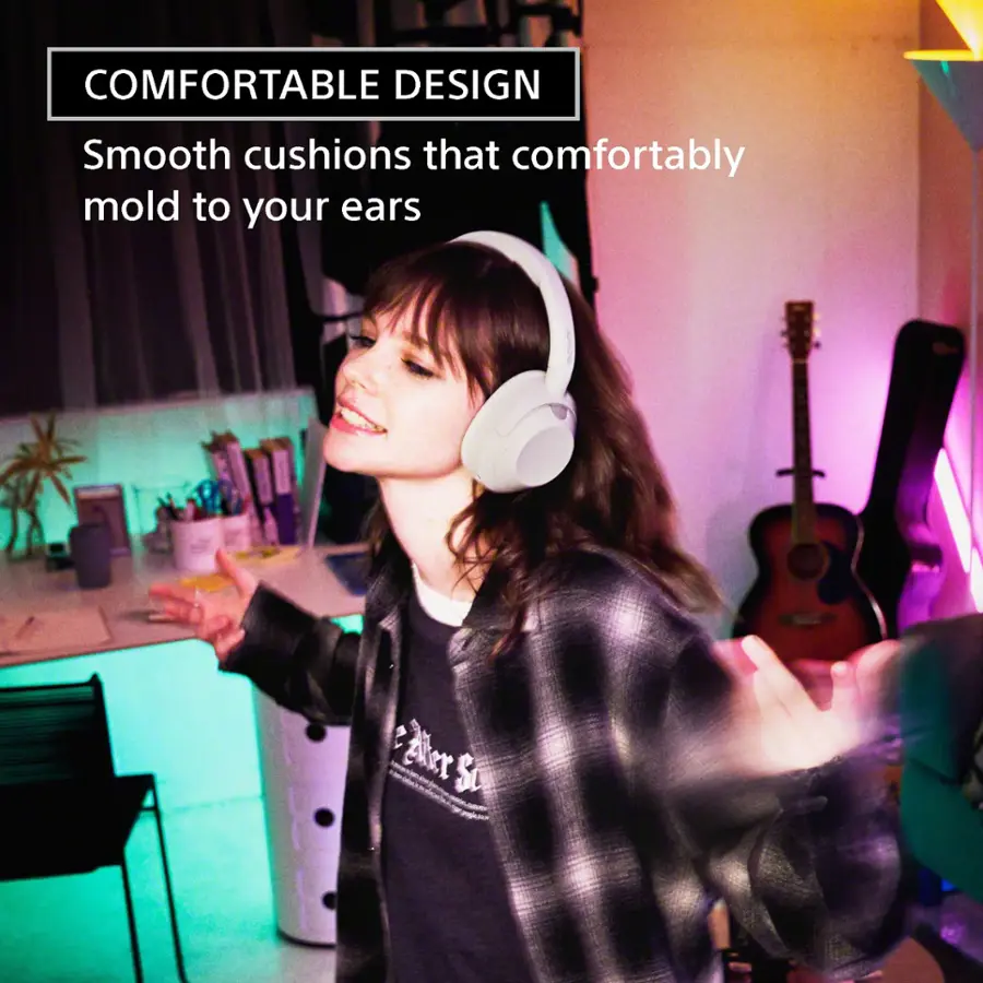 The image features a woman wearing headphones and standing in a room with a guitar. She appears to be enjoying herself and possibly singing. The headphones are described as having "smooth cushions that comfortably mold to your ears." The woman is also wearing a plaid shirt, adding to the overall visual appeal of the scene.