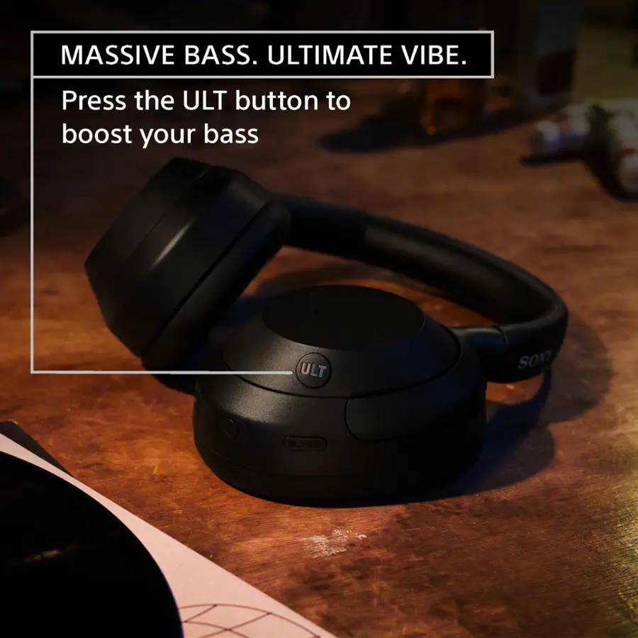 The image features a pair of black headphones with a large bass speaker on a table. The headphones are designed to provide an ultimate vibe with massive bass. To boost the bass, the user can press the ULT button. The headphones are placed on a table, surrounded by various objects such as a bottle, a cup, and a book.