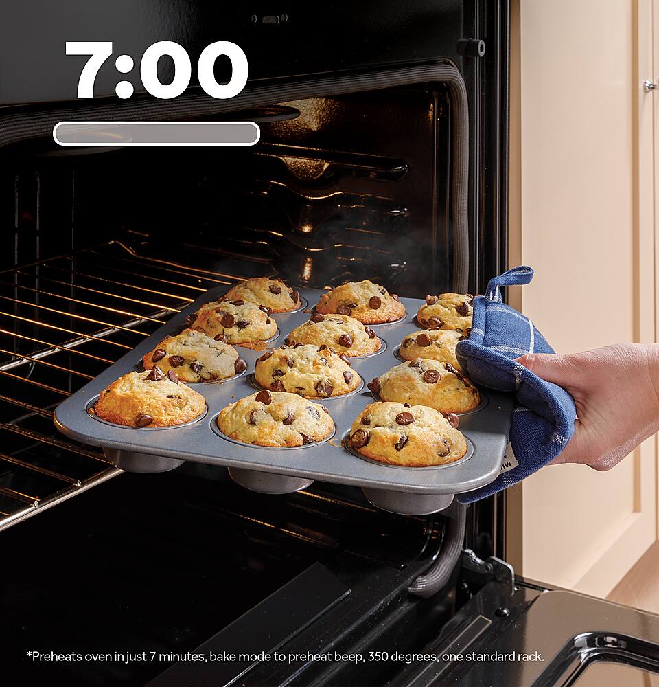 The image shows a person holding a tray of muffins that have just come out of an oven. The oven is open, and the muffins are placed on a cooling rack. The person is likely in the process of removing the muffins from the oven, as they are now ready to be served. The oven is a standard oven with a standard rack, and it preheats in just 7 minutes, making it a convenient and efficient appliance for baking.