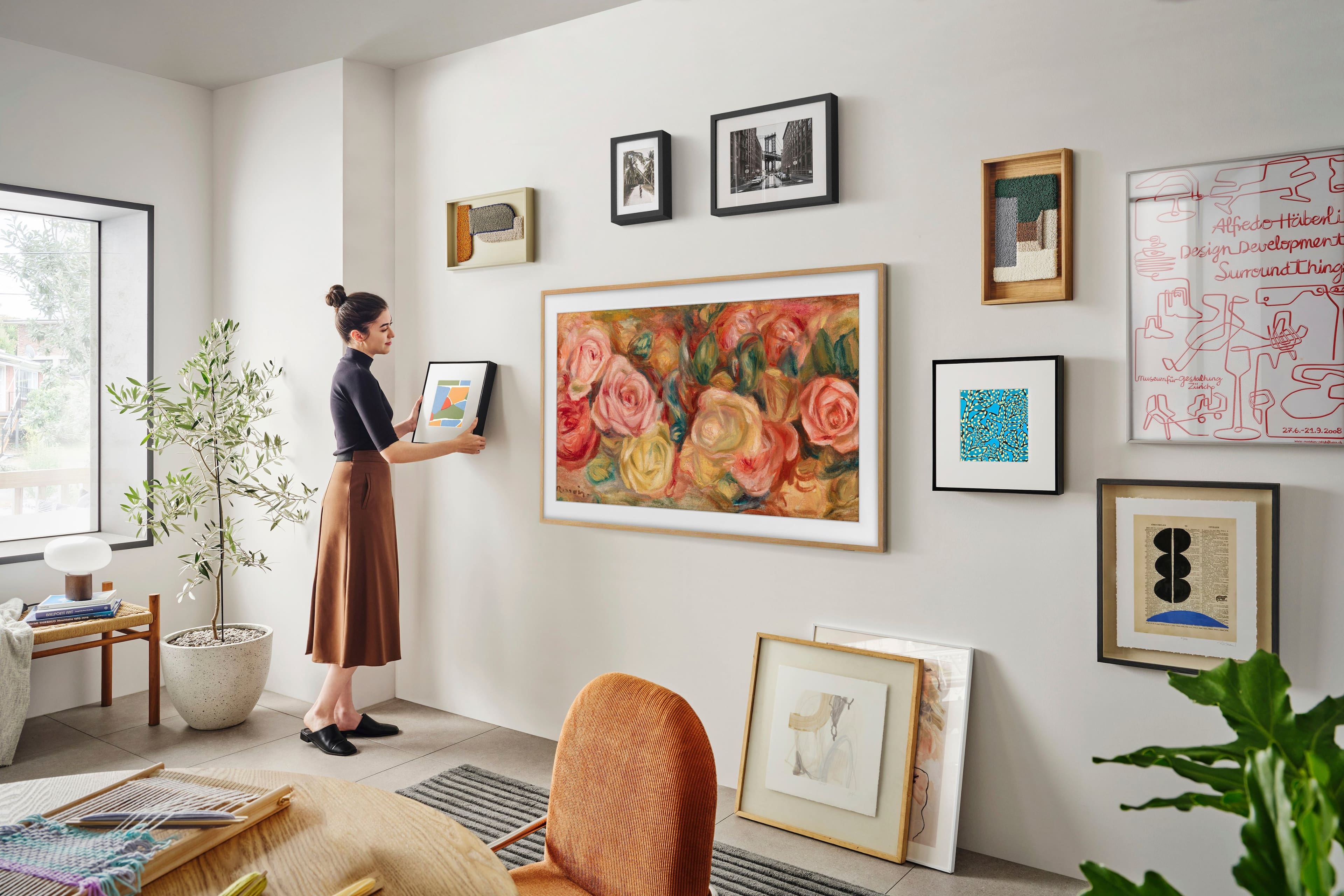 The image shows a woman standing in a living room, holding a picture frame. She is surrounded by various artworks, including paintings and photographs, which are displayed on the walls. The room also features a dining table, a chair, and a potted plant. The woman appears to be in the process of hanging or arranging the artwork on the wall.