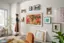 The image shows a woman standing in a living room, holding a picture frame. She is surrounded by various artworks, including paintings and photographs, which are displayed on the walls. The room also features a dining table, a chair, and a potted plant. The woman appears to be in the process of hanging or arranging the artwork on the wall.