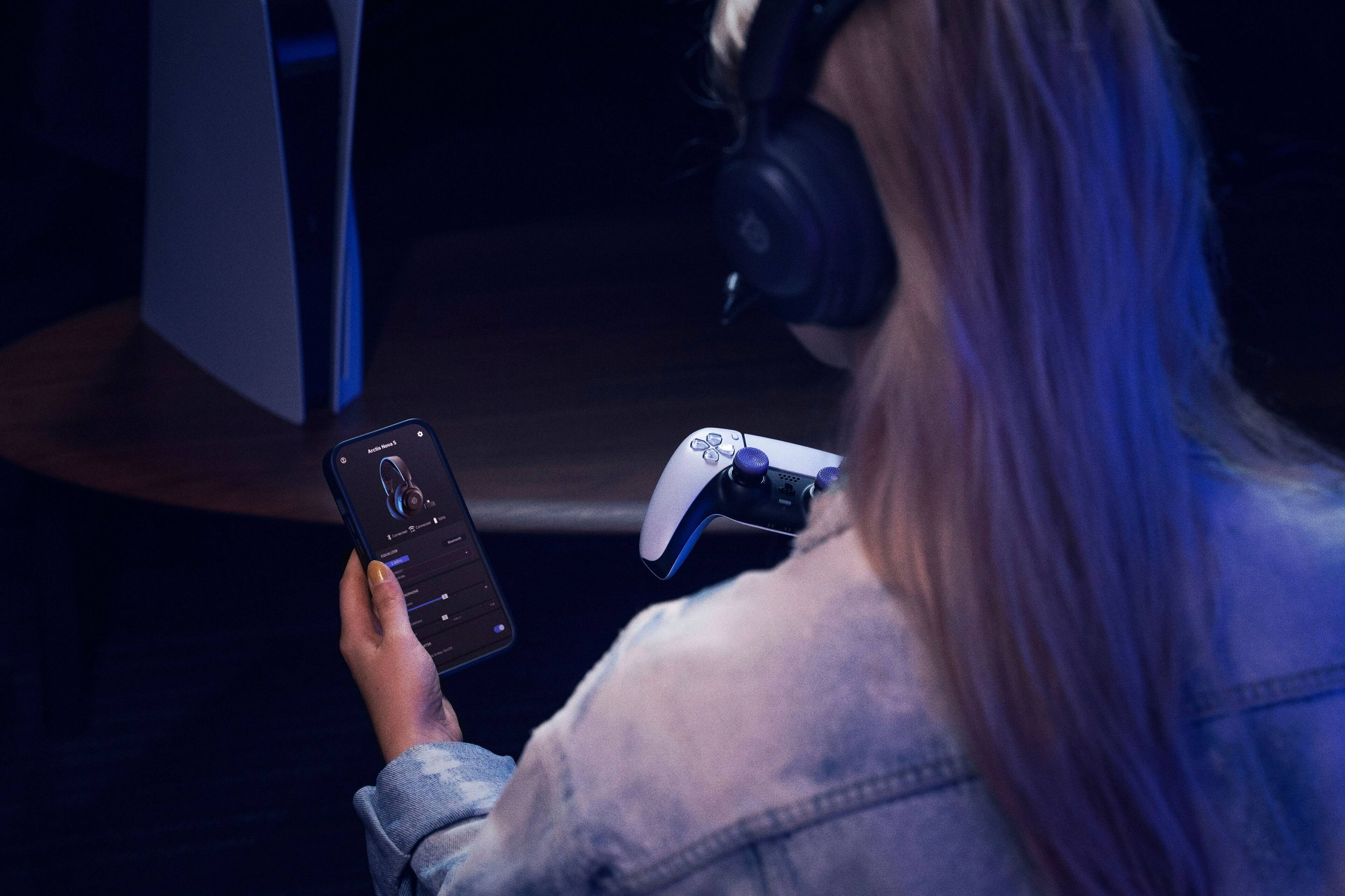 A woman is sitting in a dark room, holding a cell phone and a game controller. She is wearing headphones and appears to be playing a video game. The image captures her focused attention on the game, as she enjoys her gaming session.