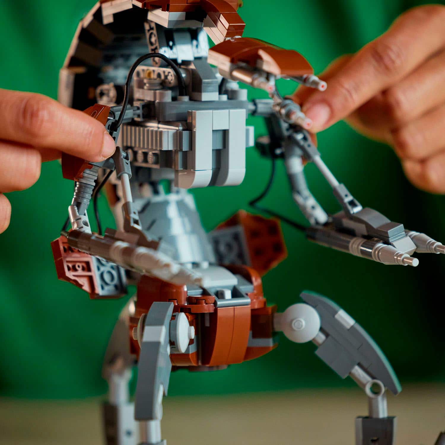 The image shows a person holding a small robot made of Legos. The robot is a unique creation, with a combination of brown and white colors. The person is pointing to the robot, possibly showing off their creation or explaining its features. The robot is positioned on a table, and the person's hand is visible in the foreground, indicating their interaction with the robot.