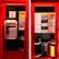 The image features a red telephone booth with a payphone inside. The payphone has a variety of stickers on it, including one that says "Here." The booth is located in a city, and there are several signs posted around it. One of the signs is a large sign that says "Big City Sightseeing." Another sign is a smaller one that says "Please be brief." The booth also has a coin slot for paying for the phone call.