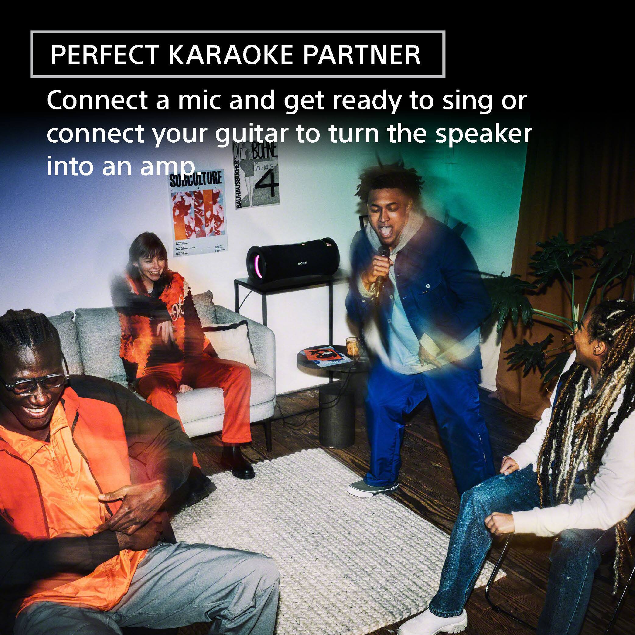 The image shows a group of people gathered in a living room, enjoying a karaoke session. There are two men and a woman singing into a microphone, while another man is playing a guitar, connecting it to the speaker to turn it into an amp. The room is furnished with a couch, a chair, and a potted plant, creating a cozy and comfortable atmosphere for the karaoke party.