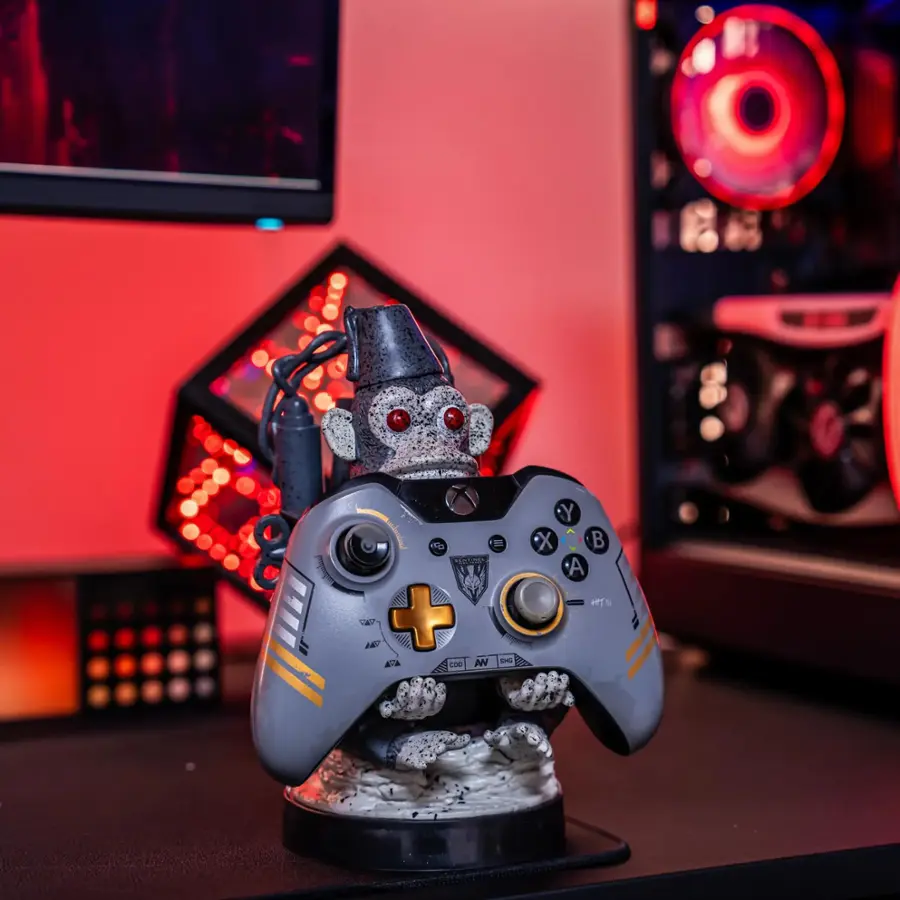 The image features a small action figure of a monkey holding a video game controller. The monkey is sitting on a desk, and the controller is placed in its hands. The scene appears to be set up for a gaming session or a display of gaming-related items.