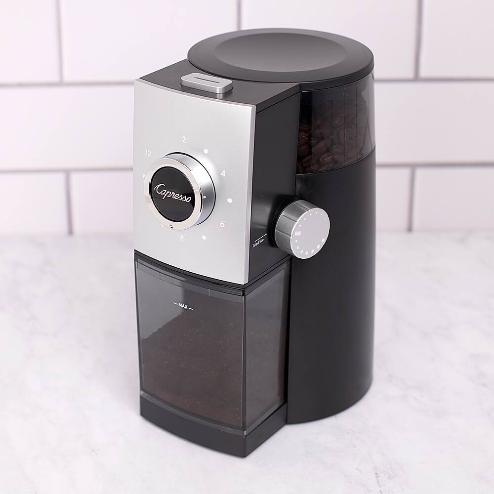 The image features a Capresso coffee maker with a glass container of coffee beans. The coffee maker is black and silver in color and is placed on a white countertop. The beans are visible in the container, ready to be used for brewing coffee.