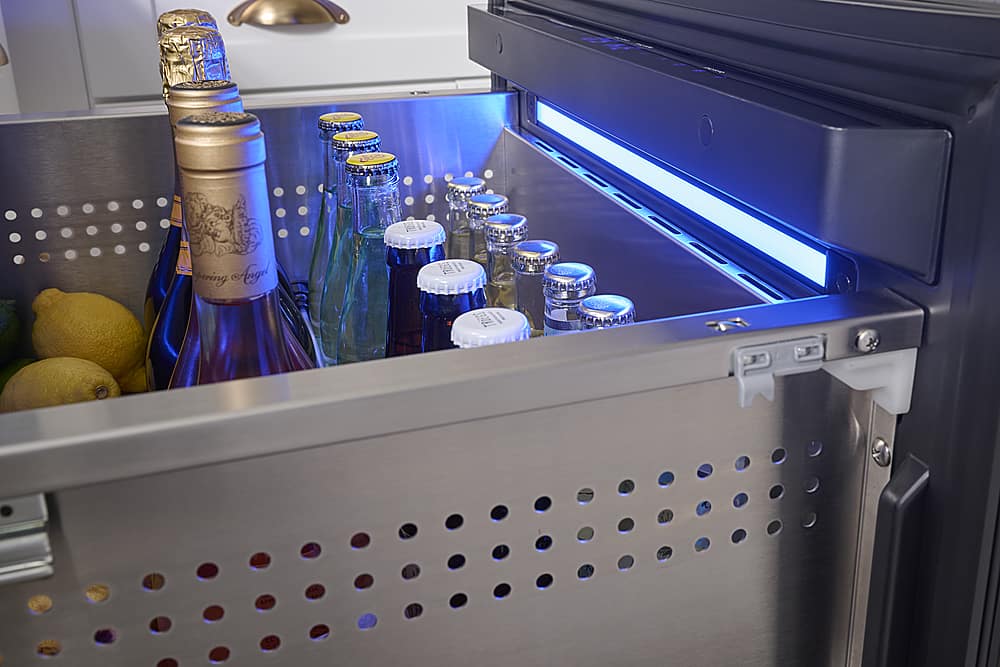 The image shows a refrigerator with a drawer filled with various bottles of alcohol, including wine and beer. The bottles are arranged in a neat and organized manner, with some bottles placed on the left side of the drawer and others on the right side. The refrigerator is also equipped with a blue light, which illuminates the bottles and adds a decorative touch to the scene. The presence of multiple bottles suggests that the refrigerator is well-stocked and ready to serve drinks to guests or for personal enjoyment.