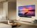 The image features a large flat screen TV mounted on a wall in a living room. The TV is displaying a beautiful sunset scene. The room is furnished with a couch and a coffee table, which has several books on it. There is also a vase placed in the room, adding to the overall aesthetic of the space.