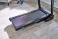 The image features a black treadmill with a blue handle on the side, placed on a grey carpet. The treadmill is part of a Stride Sport machine, which is designed for exercise and fitness purposes. The machine is placed in a room with a couch visible in the background.