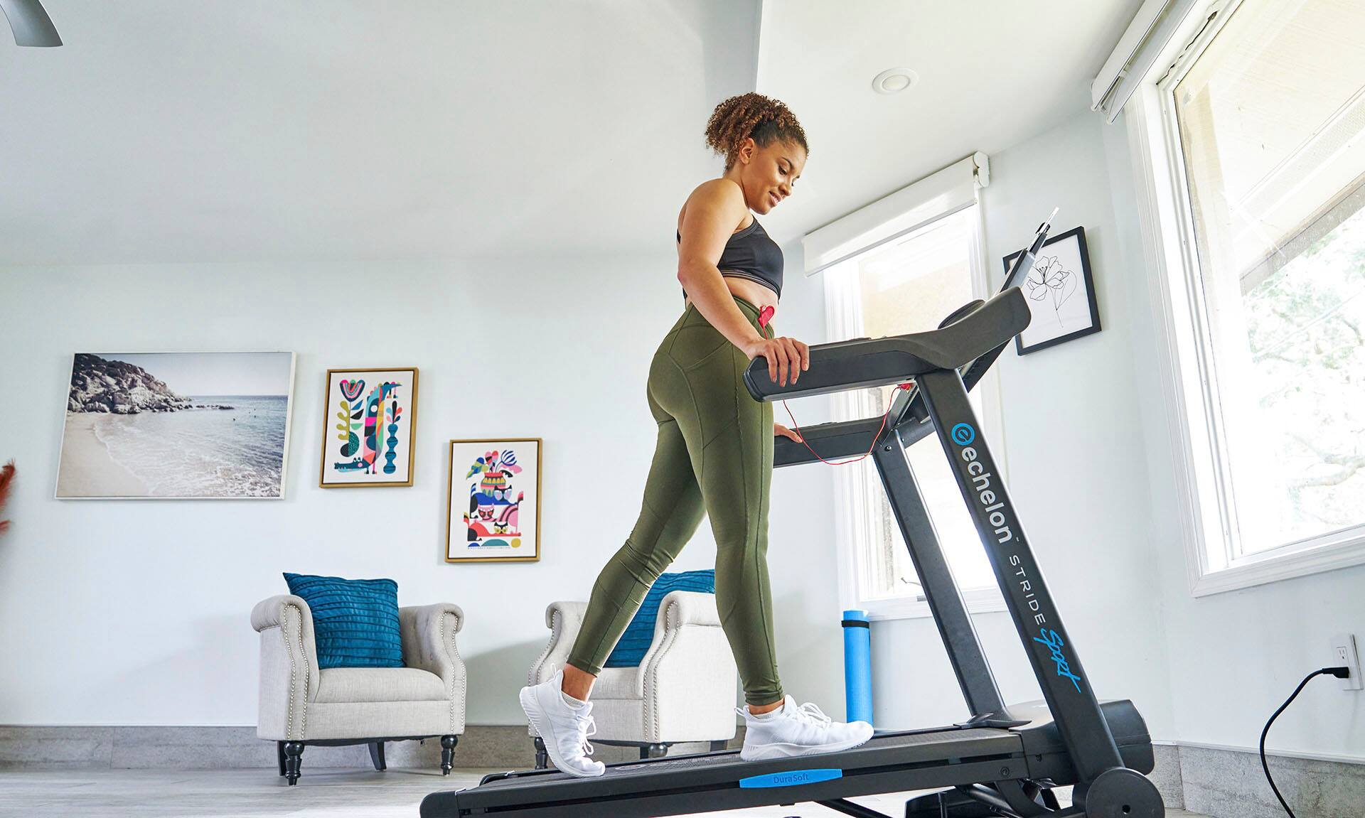 The woman in the image is using an Echelon Stride machine, which is a piece of exercise equipment designed for indoor use. The machine is placed in a living room, and the woman is wearing a black shirt and green pants while standing on the machine. The Echelon Stride is a durable and soft exercise machine that provides a comfortable and effective workout experience.