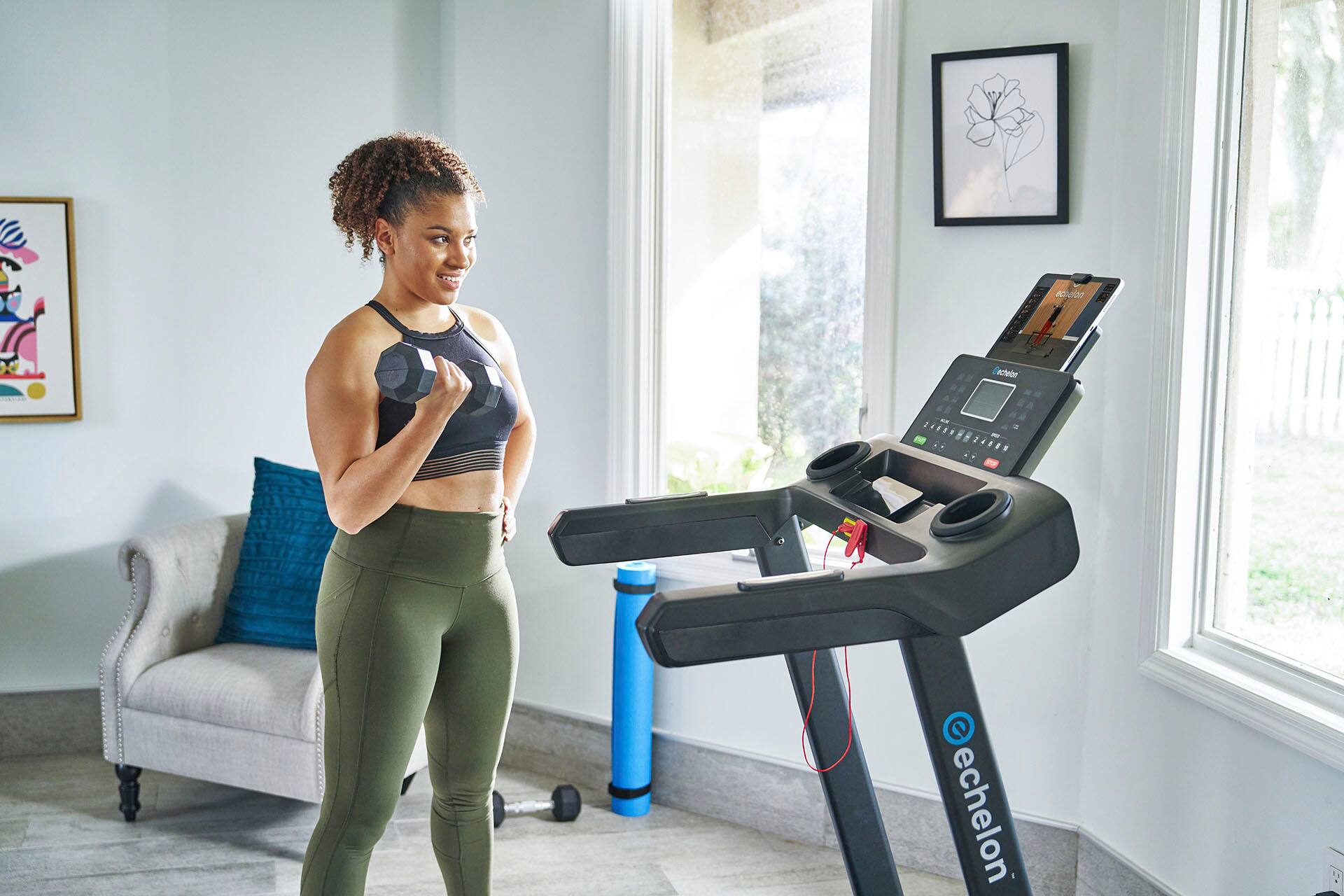 The woman is standing in a room, wearing a black tank top and green leggings. She is using a stationary bike, which has a screen in front of her. She is smiling and appears to be enjoying her workout. The room has a couch in the background, and there is a picture hanging on the wall.