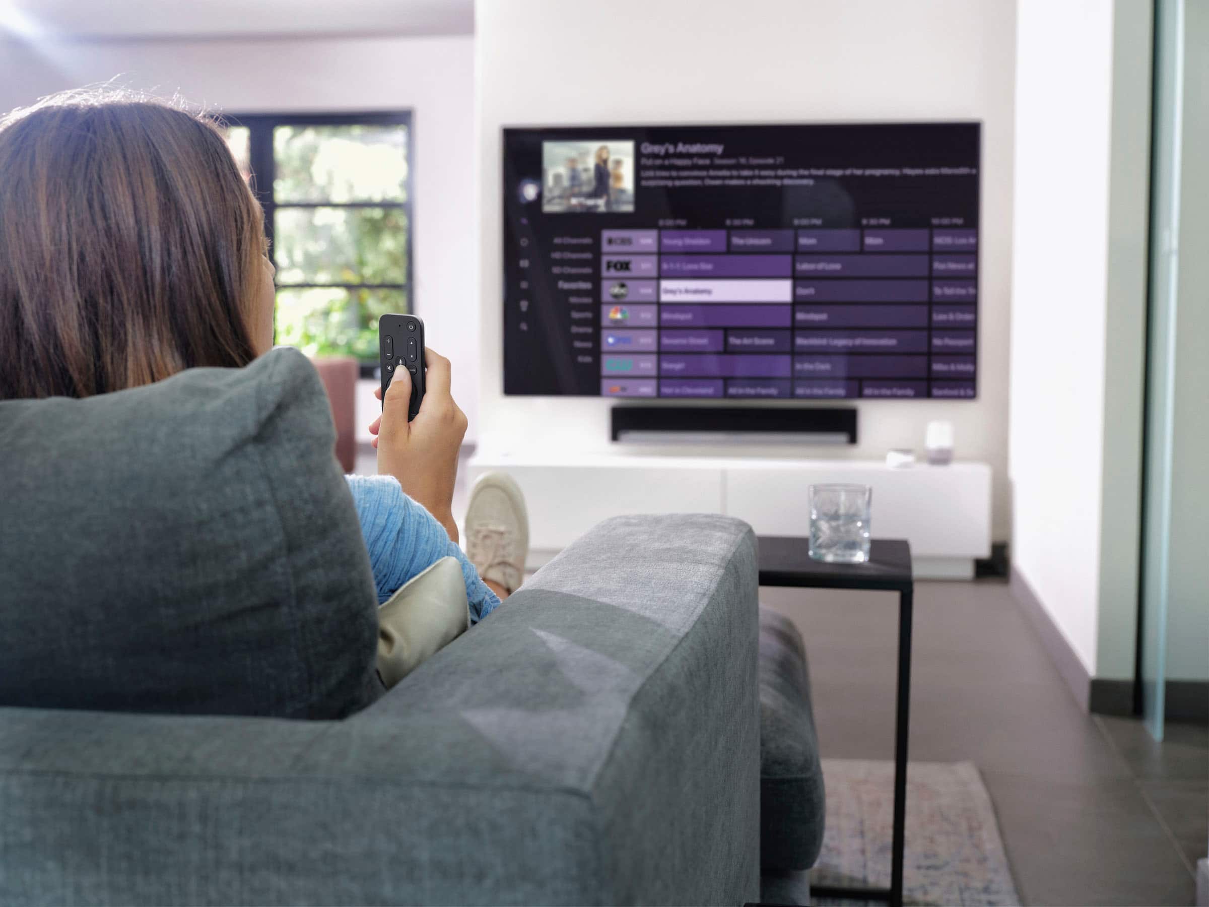 The woman is sitting on a couch, holding a remote control in her hand, and watching a television. The TV is displaying the show "Grey's Anatomy" on the FOX channel.