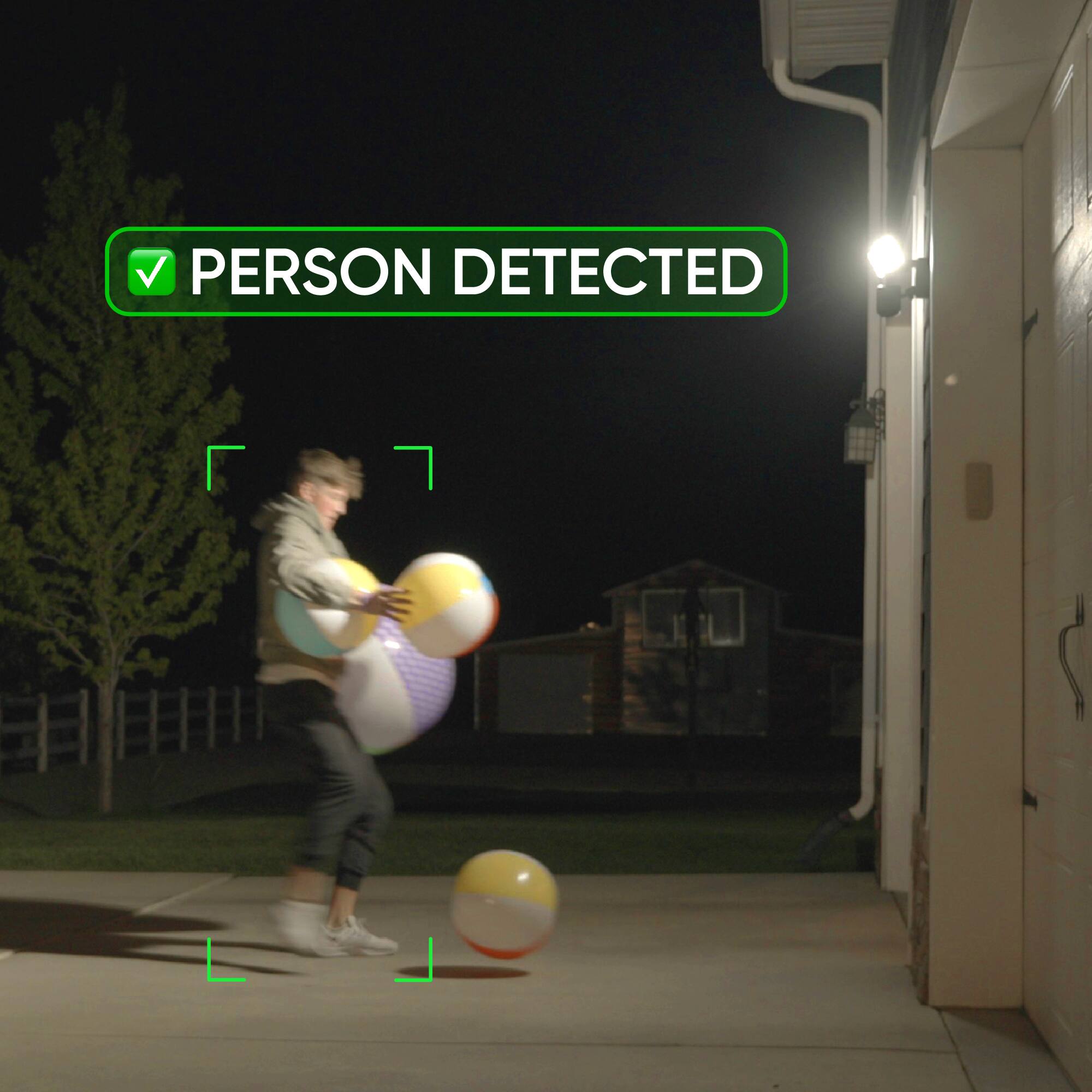 In the image, a person is standing on a driveway, holding two large beach balls. The scene is captured at night, and there is a green "Person Detected" sign on the screen. The person appears to be playing with the beach balls, possibly enjoying a game or simply having fun. The presence of the "Person Detected" sign suggests that there might be a security system in place to monitor the area.