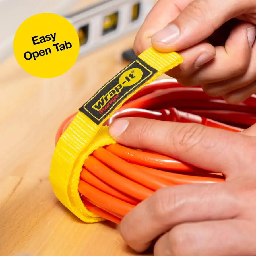 The image features a person holding a yellow Wrap-it cable tie in their hand. The cable tie is being used to secure a cord, and the person is wrapping the tie around the cord. The scene takes place on a wooden table, where the person is focused on securing the cord with the Wrap-it cable tie.