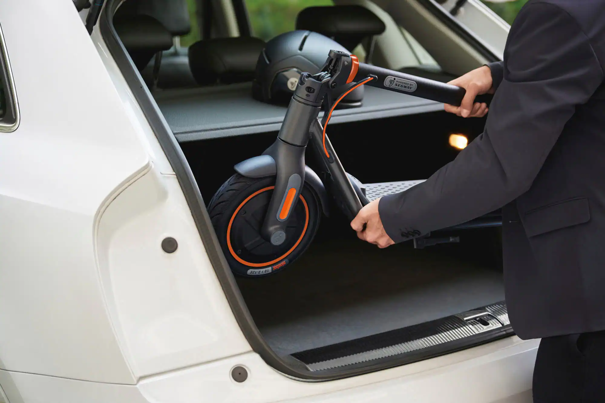 Zoom in on The image shows a man in a suit holding a helmet and a Segway C-100 scooter. He is in the process of putting the helmet into the back of a car. The man is wearing a black suit and is standing next to the car. The scene captures the moment of the man preparing to transport the helmet and scooter to their destination.