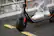The image features a person wearing white shoes and riding a Ninebot scooter. The scooter is black and has a yellow line on the ground. The person is standing on the scooter, which is parked on the sidewalk.