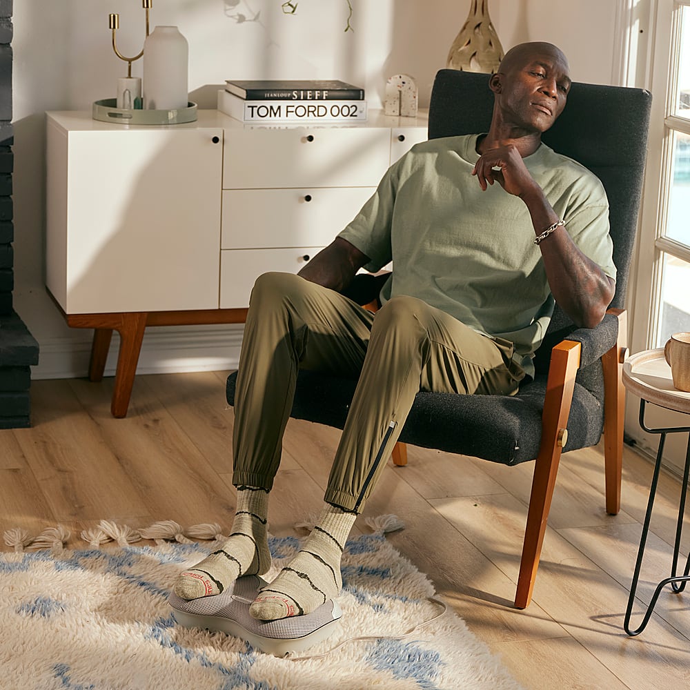 The man is sitting in a chair with his legs crossed, wearing a green shirt and socks. He is relaxed and appears to be resting or taking a break. The room he is in has a white rug, and there is a book nearby. The man is also wearing a watch on his wrist.