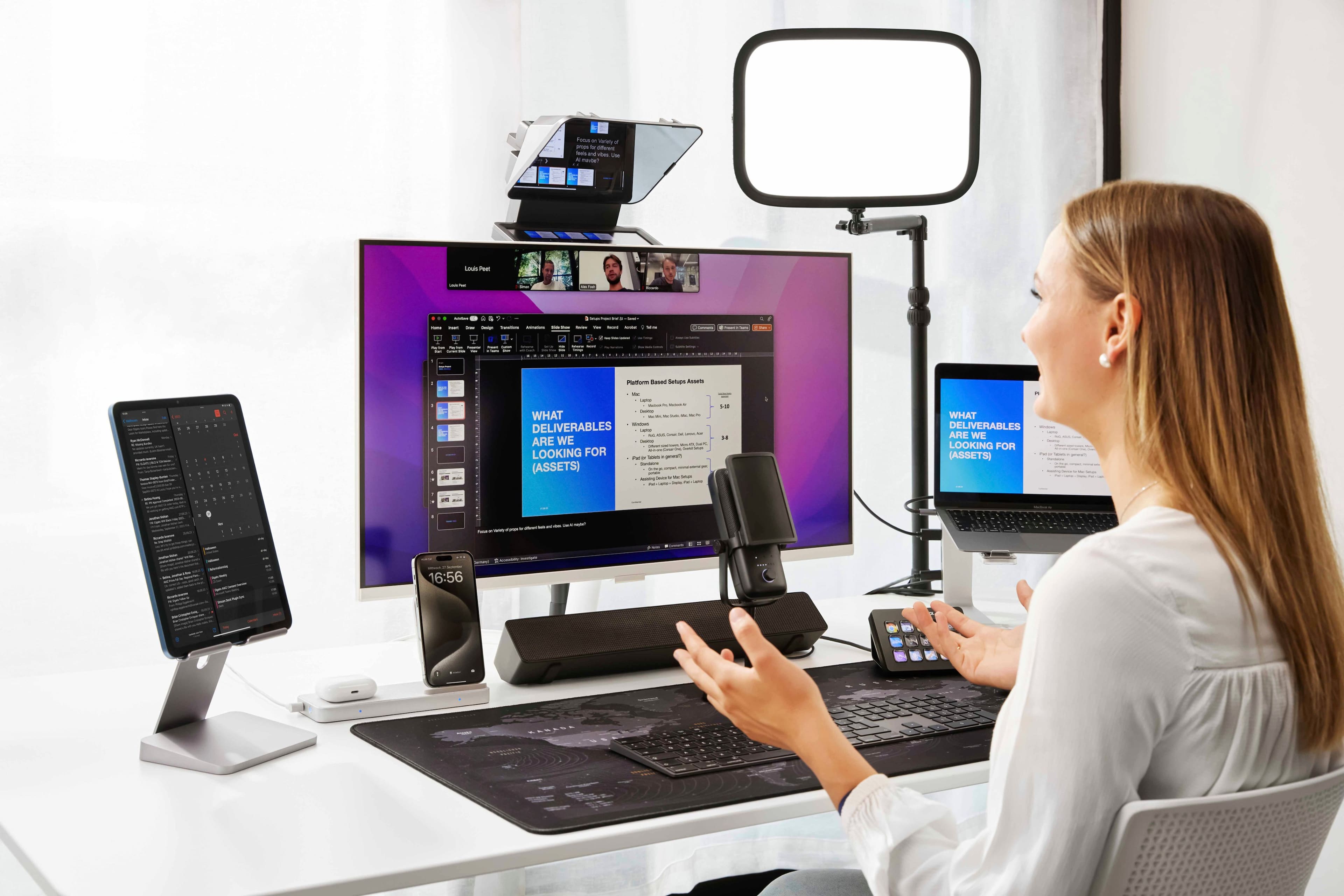 The image features a woman sitting at a desk with a variety of electronic devices. There is a laptop, a desktop computer with a monitor, a cell phone, and a microphone. The woman appears to be engaged in a video conference or working on a project that requires multiple screens and devices. The scene captures the modern workspace, showcasing the importance of technology and communication tools in today's professional environment.