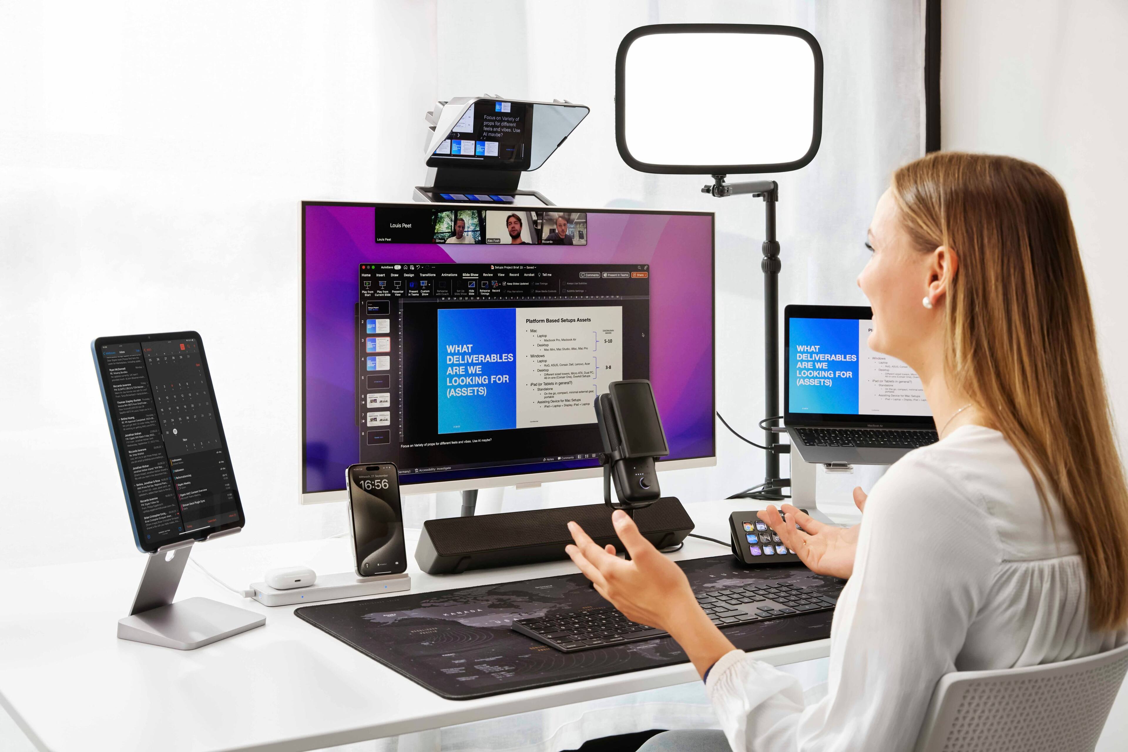 The image features a woman sitting at a desk with a variety of electronic devices. There is a laptop, a desktop computer with a monitor, a cell phone, and a microphone. The woman appears to be engaged in a video conference or working on a project that requires multiple screens and devices. The scene captures the modern workspace, showcasing the importance of technology and communication tools in today's professional environment.