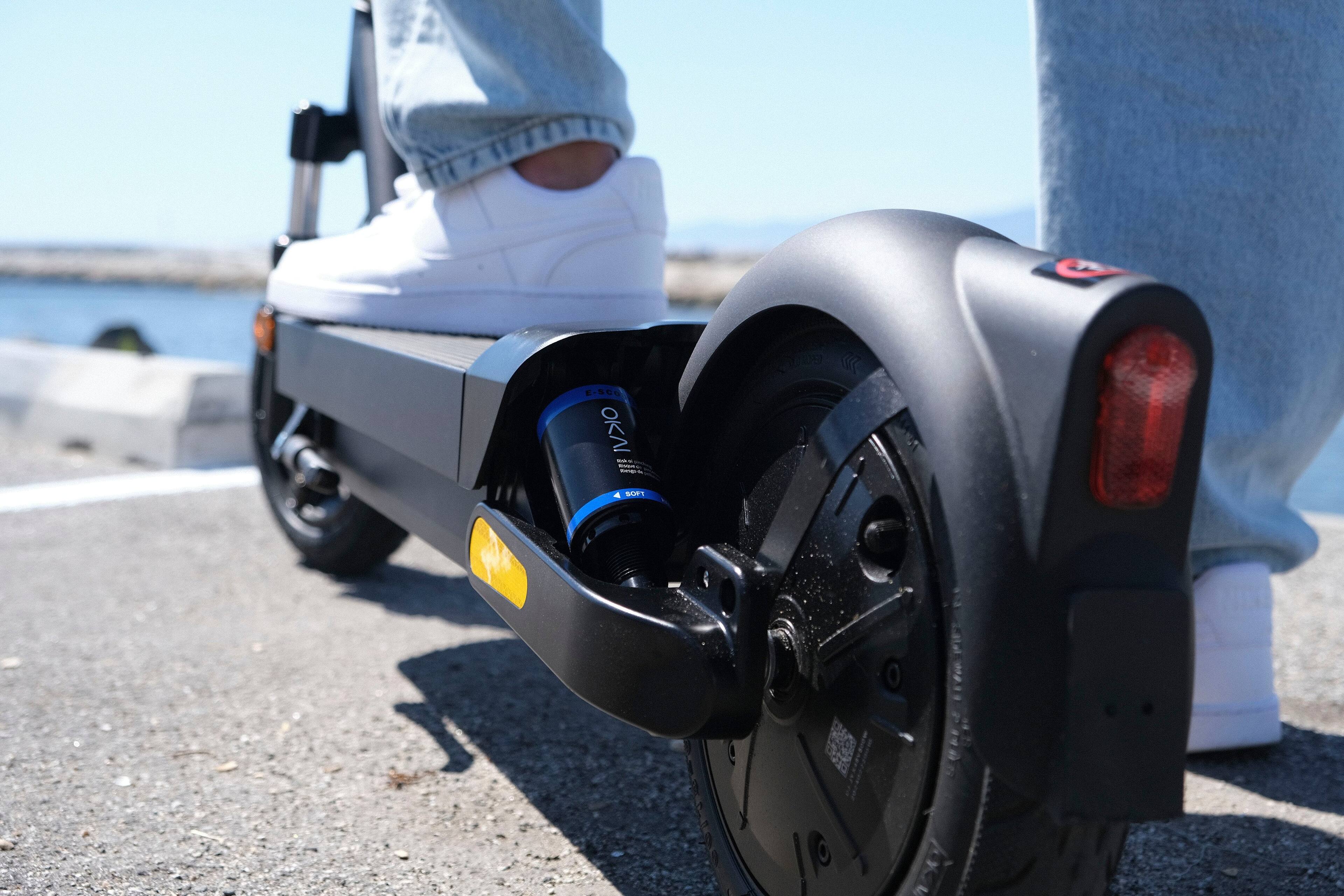 The image shows a person standing on a scooter, with their feet on the footrests. The scooter has a large wheel and a black wheel guard. The person is wearing white shoes, and the scooter is located on a sidewalk near a body of water. The scene appears to be taking place during the day.