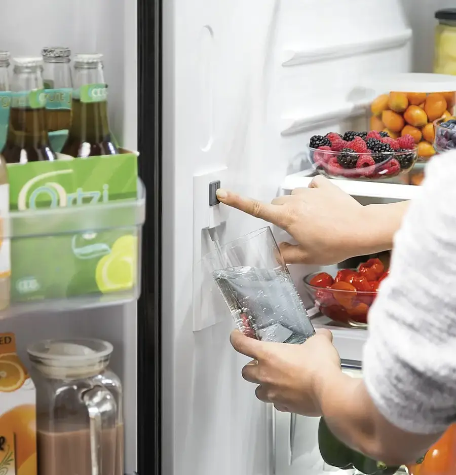 The image shows a person reaching into a refrigerator to grab a bottle of water. The refrigerator is filled with various food items, including fruits and vegetables. The person is holding the water bottle in their hand, and the refrigerator door is open.