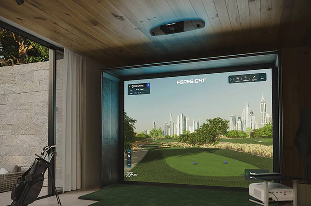 The image shows a golf course being displayed on a large screen, likely in a living room or a golf simulator. The screen is showing a golf course with a golf ball in motion, indicating that it is a golf simulation game. The screen is placed in front of a couch, and there is a golf club visible in the scene. The room appears to be designed for a comfortable and immersive golfing experience.