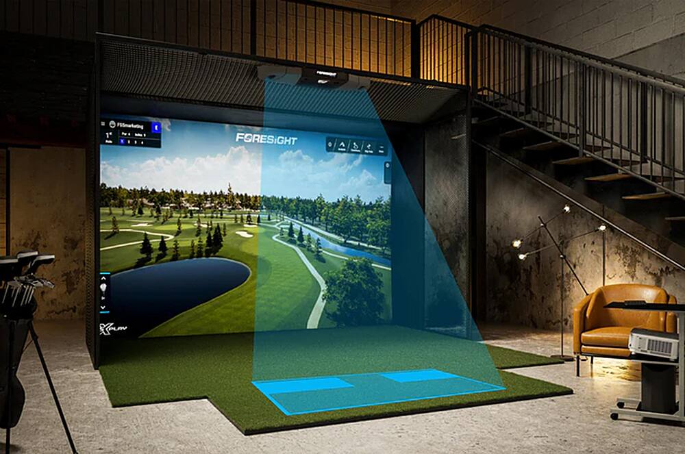 The image features a large screen displaying a golf course, likely a ForeSight golf simulator. The screen is located in a dark room, possibly a basement, and is surrounded by a few chairs. The room also has a staircase leading up to the next level.