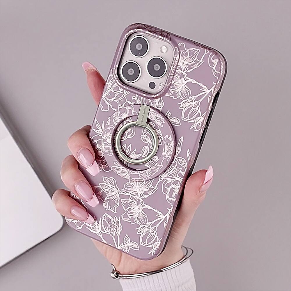 The image shows a person holding a cell phone in their hand. The phone is covered with a purple case featuring a flower design. The person's nails are painted with a pink color, and they are wearing a white sweater. The cell phone is held in a way that the flower design on the case is visible, and the person's hand is positioned in a way that the phone is easily visible.
