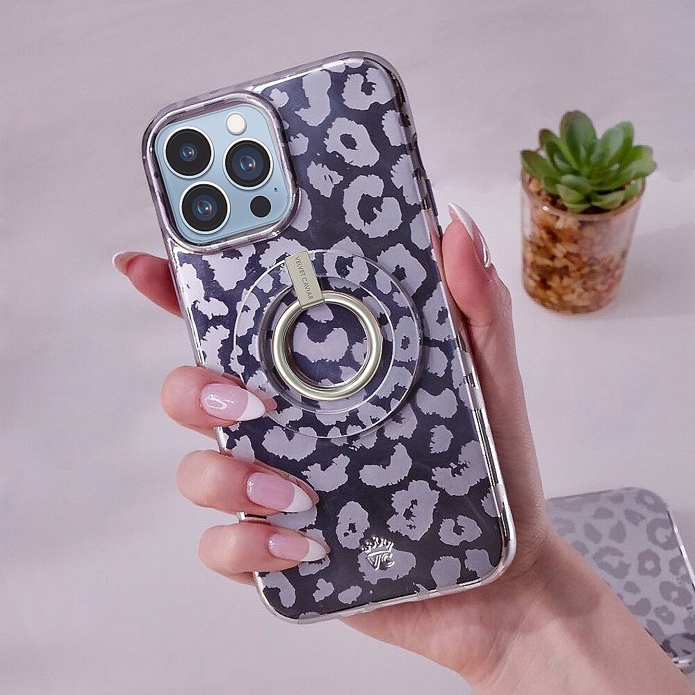 The image shows a person holding a cell phone with a leopard print case. The case has a ring on it, and the phone is being held in front of the person's hand. The person's nails are painted, and they are wearing a ring on their finger. The scene also includes a potted plant in the background.