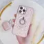 The image shows a person holding a pink cell phone in their hand. The phone is covered with a pink case, and it appears to be a smartphone. The person is also holding a small pink purse, which is placed next to the cell phone. The scene takes place on a table, and there is a gold ring nearby.