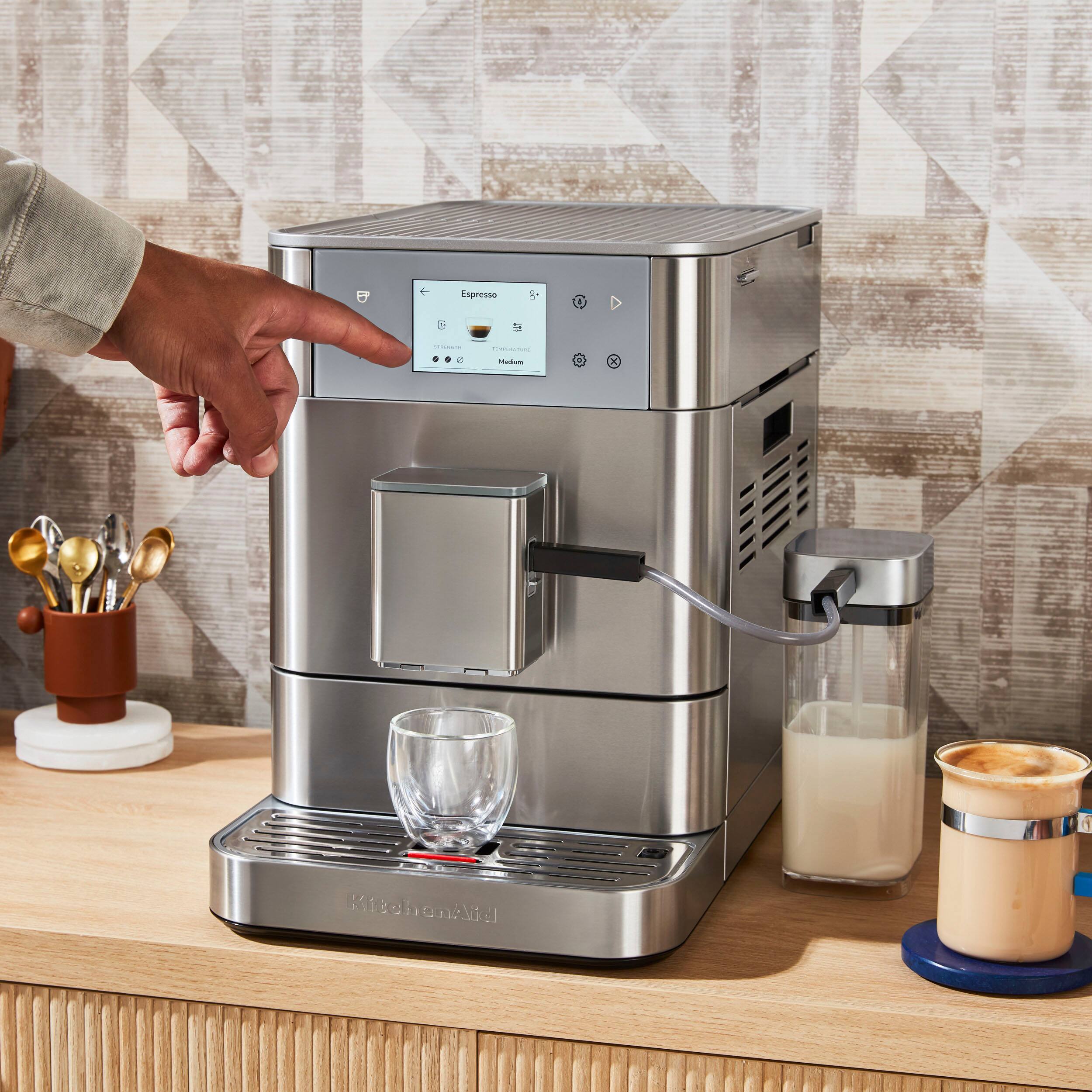 The image features a person pointing at a coffee maker, which is sitting on a counter. The coffee maker is connected to a glass container and a milk container, indicating that it is a fully automatic coffee machine. The person is likely demonstrating how to use the machine or showing its features. The scene also includes a cup and a spoon nearby, suggesting that the coffee maker is ready to be used.