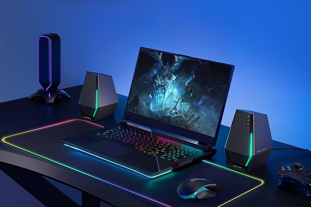The image features a laptop computer sitting on a desk with a keyboard and mouse. The laptop is open and turned on, displaying a video game on its screen. There are also two speakers on the desk, one on each side of the laptop. The desk is illuminated by a blue light, creating a gaming setup with a neon glow.