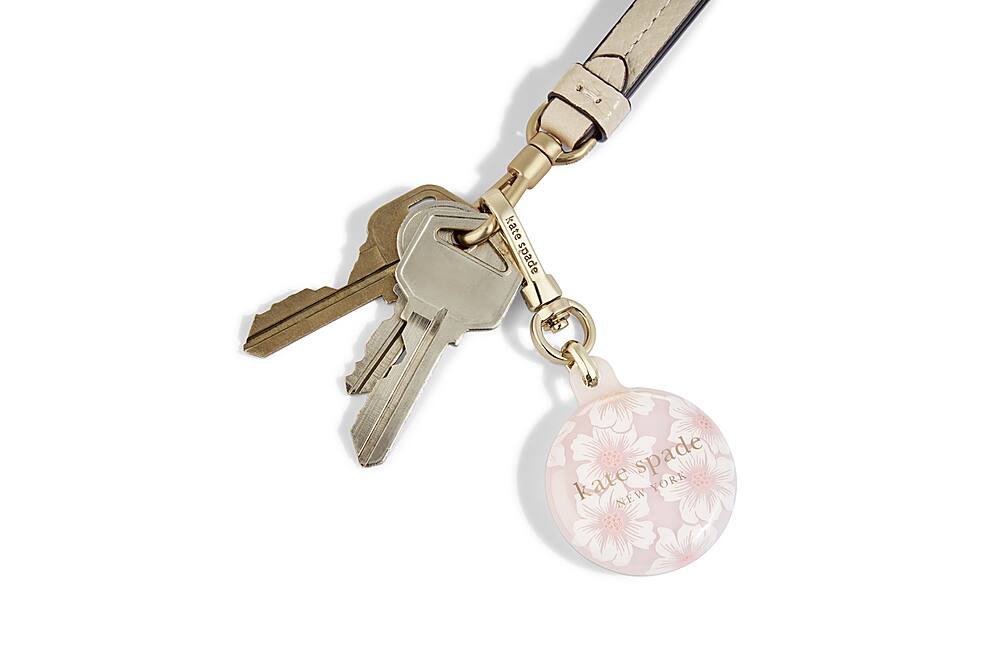 The image features a keychain with a pink flower charm and two keys. The keychain is hanging from a black strap. The charm is attached to the keys, making it a functional and stylish accessory. The keychain is designed by Kate Spade New York, a well-known fashion brand.