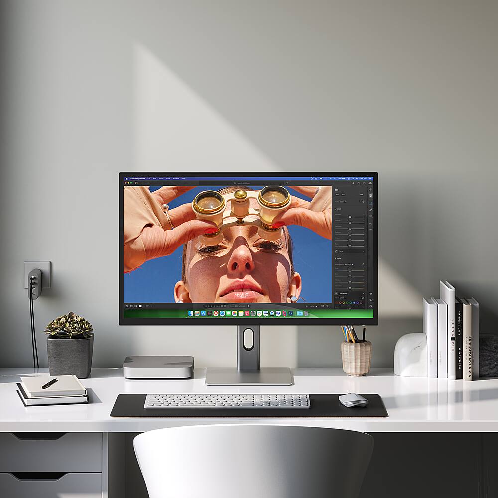 The image shows a desk with a computer monitor, keyboard, and mouse. The computer monitor displays a picture of a woman wearing glasses. There are also several books on the desk, and a potted plant is placed nearby. The desk is situated in front of a window, providing natural light to the workspace.