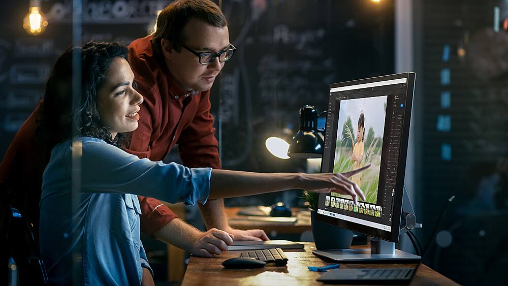 The image shows a man and a woman working together on a laptop. The man is sitting at the desk, while the woman is standing behind him. The woman is pointing at the laptop screen, which is displaying a picture of a dog. The man is also pointing at the screen, possibly discussing the content or providing input. There are two keyboards on the desk, one in front of the man and another in front of the woman. Additionally, there is a cup placed on the desk, likely for refreshments during their work session.