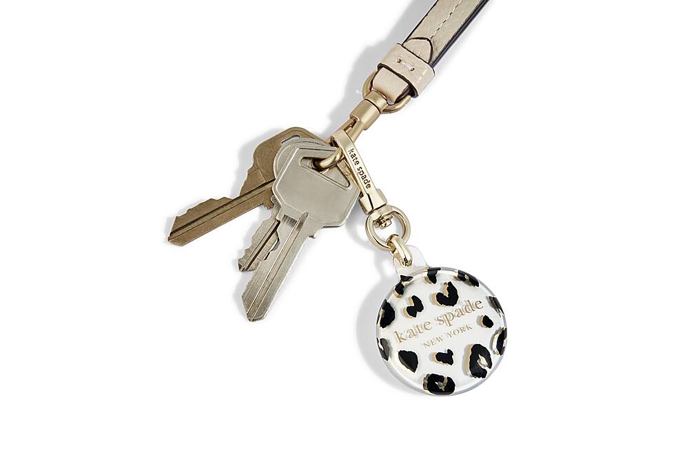 The image features a keychain with a key ring and a round charm. The charm has a leopard print design, and the key ring is made of gold. The keychain is attached to a purse strap, which is also made of gold. The purse strap is hanging down, and the keychain is prominently displayed in the foreground.