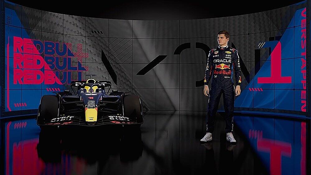 The image features a man standing in front of a Red Bull racing car. The man is wearing a blue and red suit, which matches the colors of the car. The car is prominently displayed in the scene, with the Red Bull logo visible on the side. The man appears to be the driver of the car, showcasing his racing suit and the car as part of a promotional event or a racing competition.