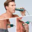 The image shows a man using a green electric shaver to shave his chest hair. He is holding the shaver close to his chest and appears to be focused on the task. The man is shirtless, which allows for a clear view of his chest hair. The shaver is a handheld device, and the man is using it to maintain his personal grooming.