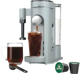The image features a coffee machine, which is likely a Ninja Coffee Bar, sitting on a counter. The coffee machine is filled with coffee and has a cup of coffee on the side. There is also a spoon and a cup of tea nearby. The scene suggests a cozy and inviting atmosphere for enjoying a hot beverage.