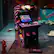 The image features a purple and black arcade machine with a large screen, likely a video game console. The machine is placed on a dirt floor, and there is a fire hydrant nearby. The scene also includes a few other objects, such as a cup, a bottle, and a book.