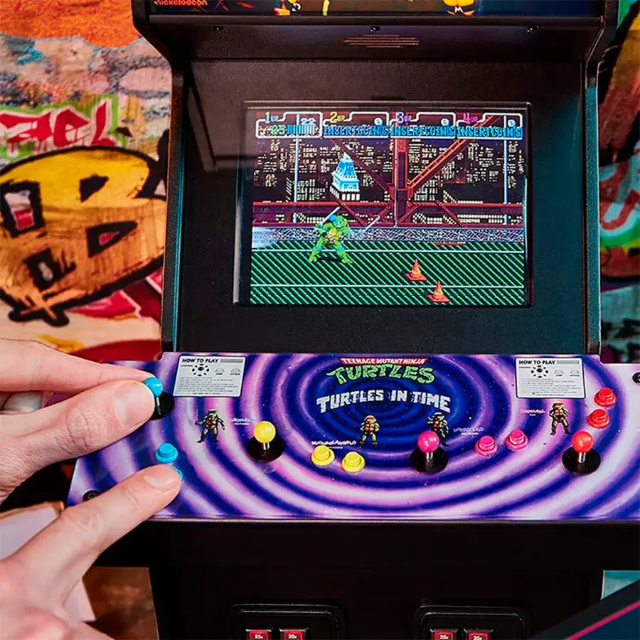 The image shows a person playing a video game, specifically the Teenage Mutant Ninja Turtles game. The person is holding a joystick, and the game is displayed on a screen. The game is set in a futuristic city, and the player is controlling a character as they navigate through the game. The game appears to be an arcade-style game, with buttons and a joystick for input.