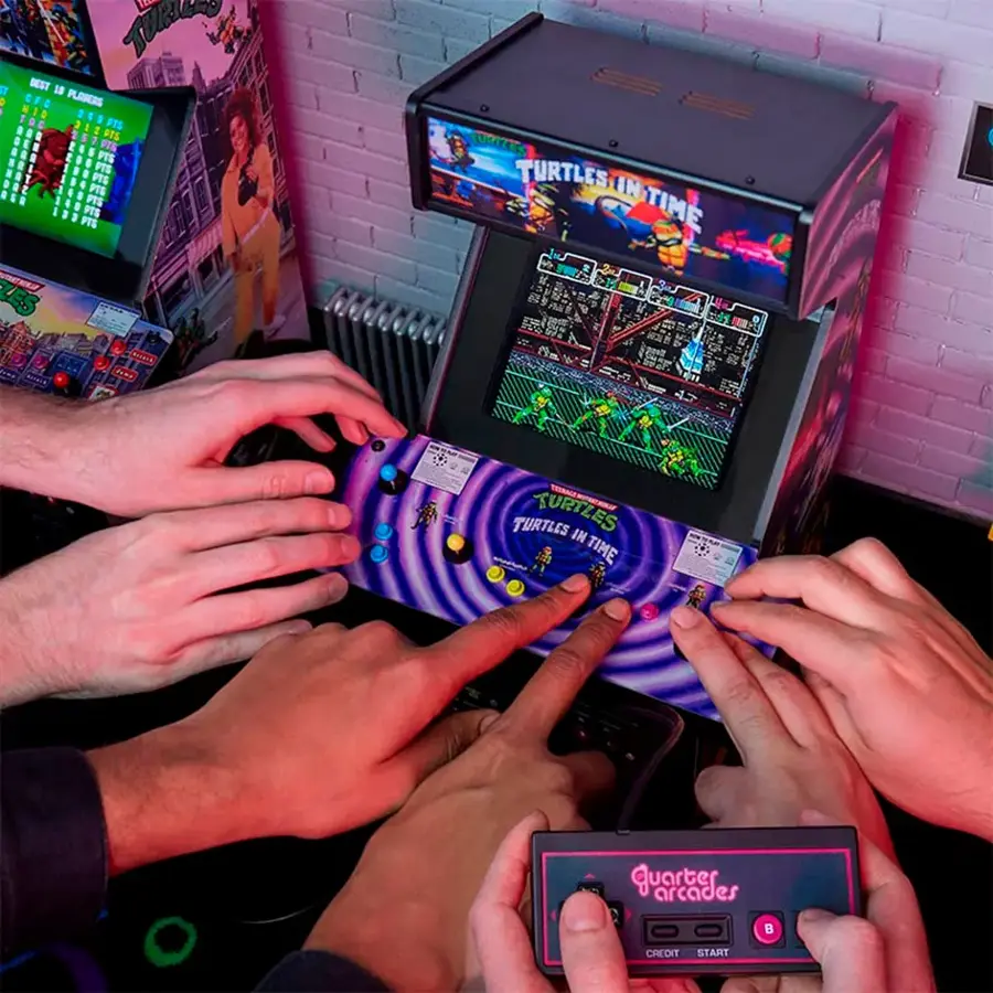 The image shows a group of people playing a video game, specifically Turtles in Time, on an arcade machine. There are several hands visible on the machine, with some people actively playing the game and others watching. The arcade machine is placed in a room with a brick wall, and there are other video game machines nearby. The game appears to be a popular choice among the group, as they gather around the machine to enjoy the gaming experience together.